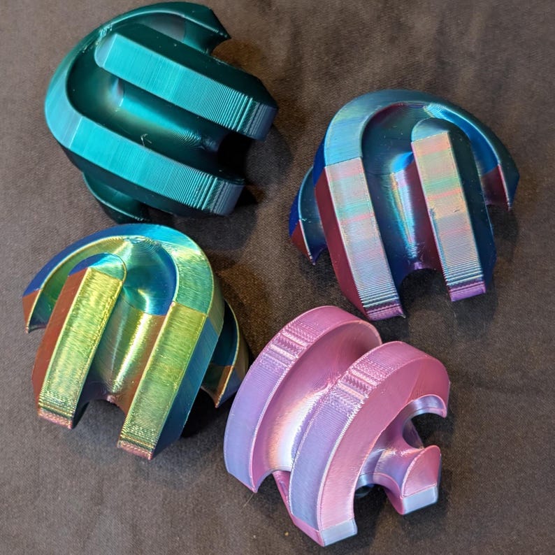 May include: Four abstract, iridescent objects with a curved, layered design. The colors shift from teal to purple and pink, creating a dynamic visual effect. The objects appear to be 3D-printed, with visible layer lines.
