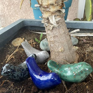 Peut inclure: Plusieurs figurines d'oiseaux en céramique de différentes couleurs, dont bleu, vert et noir moucheté de blanc. Les oiseaux sont disposés dans un pot avec de la terre et des cailloux, près d'un tronc d'arbre.