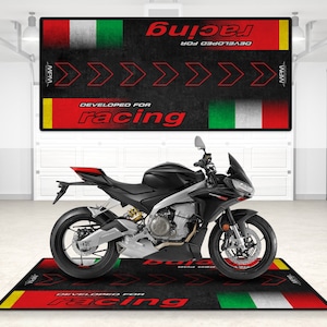 Puede incluir: Una motocicleta negra con detalles rojos está aparcada en una alfombra de suelo negra y roja con las palabras "Desarrollado para carreras" y la bandera italiana. La alfombra tiene un ribete amarillo.