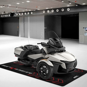 Op de afbeelding: Een wit-zwarte driewielige motorfiets met een zwarte voorruit en een zwarte stoel. De motorfiets staat op een zwarte mat met het woord "SPRINTER" in rood.