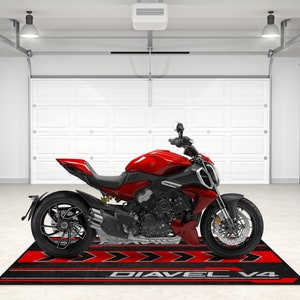 Op de afbeelding: Een rode Ducati Diavel V4 motorfiets geparkeerd in een garage op een zwart en rode motorfietsmat met de woorden "Diavel V4" erop gedrukt.
