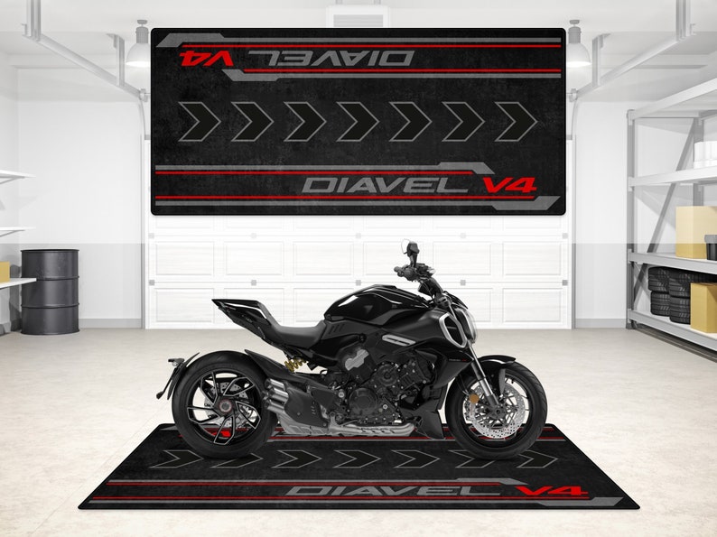 Op de afbeelding: Zwarte motorfiets met rode en grijze accenten op een zwarte en rode garagevloermat. De mat heeft de tekst "Diavel V4" erop gedrukt.