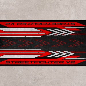 Puede incluir: Alfombrilla de rat&oacute;n para juegos de color negro y rojo con el texto "STREET FIGHTER V2" impreso en ella. El dise&ntilde;o presenta flechas blancas y rojas.