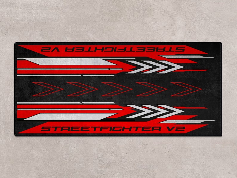 Puede incluir: Alfombrilla de rat&oacute;n para juegos de color negro y rojo con el texto "STREET FIGHTER V2" impreso en ella. El dise&ntilde;o presenta flechas blancas y rojas.
