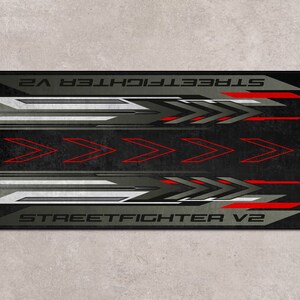 Puede incluir: Alfombrilla de rat&oacute;n negra y gris con un dise&ntilde;o rojo y blanco. El texto "STREET FIGHTER V2" est&aacute; impreso en la alfombrilla de rat&oacute;n.