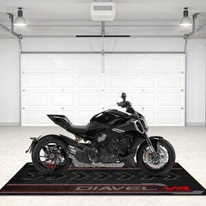 Op de afbeelding: Een zwarte motorfiets met rode accenten op de wielen en een zwarte mat met de tekst "Diavel V4" erop.