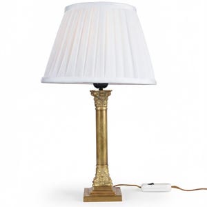 Puede incluir: Una lámpara de mesa dorada con una pantalla plisada blanca. La lámpara tiene una base en forma de columna con detalles ornamentados y una base cuadrada. La lámpara está enchufada a un cable de alimentación blanco.