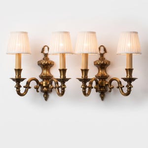May include: A pair of two-light wall sconces featuring an antique brass finish with ornate scrollwork and central decorative backplates. Each sconce is topped with two off-white pleated fabric lampshades, creating a symmetrical lighting fixture.