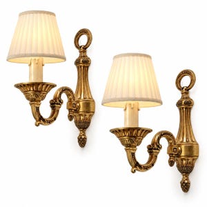 May include: A pair of wall sconces featuring gold-toned metal fixtures with ornate, fluted detailing and a ring-shaped top. Each sconce holds a single light bulb covered by a cream-coloured, pleated fabric lampshade, creating a warm, illuminated glow.