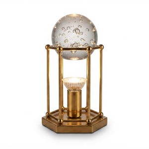 Op de afbeelding: Een tafellamp met een zeshoekige messing voet en een kooiachtig frame met verticale staven. Een heldere glazen bol met interne luchtbellen rust aan de bovenkant, verlicht door een centrale lichtbron in een cilindrische messing fitting.