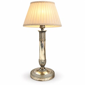 May include: A table lamp with a metallic silver-toned base featuring a textured cross-hatch pattern at the top and bottom, a leaf-like design near the base, and a pleated, off-white fabric lampshade that emits a warm, soft, diffused light.