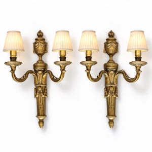 Puede incluir: Un par de apliques de pared de dos brazos con acabado en oro metálico y detalles tallados. Cada aplique tiene una columna central vertical rematada con un remate decorativo y dos brazos curvos, cada uno con una pequeña pantalla plisada de color blanco roto.