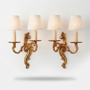 May include: A pair of wall sconces featuring ornate gold-toned metal scrollwork bases and cream-colored pleated fabric shades. Each fixture holds a single light bulb, with the metal arms extending outward to support the upright lampshades.