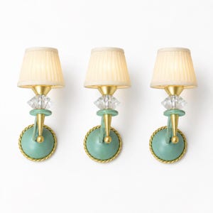 May include: Three wall sconces featuring pleated cream fabric shades, clear faceted crystal accents, and gold-toned metal hardware. Each fixture has a circular, sage green base with a decorative rope-style border and a downward-pointing arm.