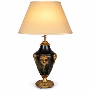 May include: A table lamp featuring a beige conical lampshade and a black urn-shaped base with a mottled finish. The base is accented with gold-coloured metal details, including ram head motifs on the sides, a fluted neck, and a circular pedestal.