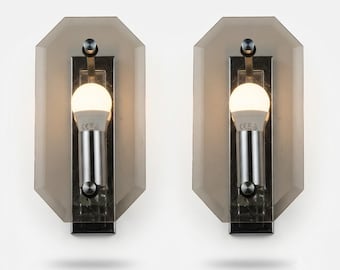 May include: A pair of wall sconces featuring octagonal smoked glass panels mounted on chrome hardware. Each fixture includes a central cylindrical chrome socket holding a single white LED bulb, secured by small circular metal fasteners.