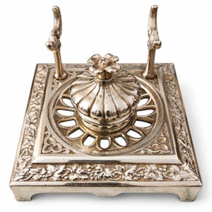 Peut inclure: Un encrier en métal argenté avec une base carrée ornée de motifs de vignes en relief. Le centre contient un pot à encre amovible, nervuré et en forme de dôme, surmonté d'un bouton floral, flanqué de deux repose-plumes verticaux.