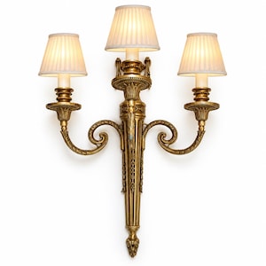 May include: A three-light wall sconce featuring an ornate gold-toned metal frame with intricate scrollwork and leaf detailing. The fixture holds three off-white, pleated fabric lampshades that emit a soft, warm glow from the internal bulbs.