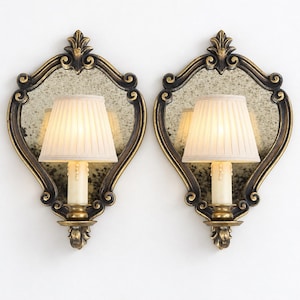 Puede incluir: Un par de apliques de pared con espejo envejecido y marco ornamentado en dorado oscuro y negro. Cada aplique tiene una lámpara con funda de vela color crema y una pantalla de tela plisada en color blanco roto.