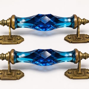 Puede incluir: Dos tiradores de cajón con un centro de cristal azul translúcido facetado con un patrón de corte de diamante. Cada tirador está asegurado por dos soportes de metal en tono latón antiguo con placas base rectangulares y orificios decorativos.