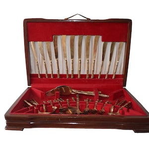 Puede incluir: Una caja de madera con un interior de terciopelo rojo, que contiene un juego de cubiertos dorados. Los cubiertos incluyen cuchillos, tenedores y cucharas.