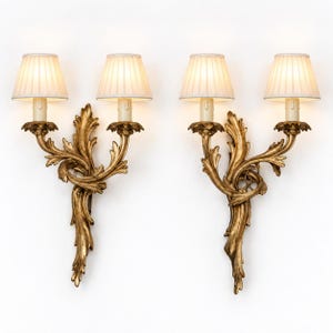 May include: A pair of two-light wall sconces featuring ornate gold-finished metalwork with scrolling acanthus leaf details. Each sconce supports two cream-colored, pleated fabric lampshades that emit a warm, soft glow from the internal bulbs.