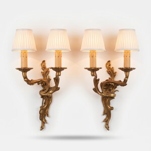 Può includere: Una coppia di applique a due luci con placche in metallo dorato decorate con volute. Ogni applique sostiene due portalampada a forma di candela, sormontati da piccoli paralumi in tessuto bianco plissettato che emettono luce calda.