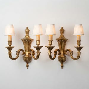 Puede incluir: Un par de apliques de pared dorados, cada uno con dos brazos y pantallas de tela blanca con nervaduras verticales. Los soportes metálicos muestran detalles ornamentales simétricos con una columna central y brazos curvos.
