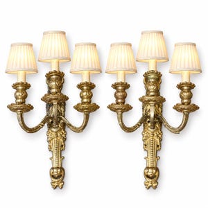 May include: A pair of gold-toned wall sconces, each featuring three arms with ornate, carved detailing and three off-white, pleated fabric lampshades. The fixtures have a vertical, decorative backplate with intricate scrollwork and floral motifs.