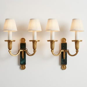 May include: A pair of two-arm wall sconces featuring gold-toned metal fixtures with ornate leaf detailing, dark green marble rectangular backplates, and four off-white pleated fabric lampshades. Each sconce has two lights with vertical stems.