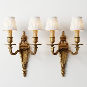 Puede incluir: Un par de apliques de pared dorados, cada uno con dos brazos que sostienen luminarias tipo vela. Cada luminaria está rematada con una pantalla de tela plisada blanca. La base de pared muestra detalles tallados y un diseño vertical.
