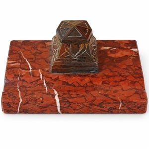 Peut inclure: Un socle rectangulaire en marbre rouge avec des veines blanches présente un encrier en métal de couleur bronze foncé centré. L'élément métallique possède un couvercle pyramidal géométrique à gradins avec des gravures linéaires.