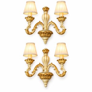 Pode incluir: Um par de arandelas de parede de duas luzes com estruturas esculpidas em tons de creme e dourado, com volutas ornamentadas e remates centrais. Cada arandela possui dois abajures de tecido plissado em tom off-white com bordas trançadas.