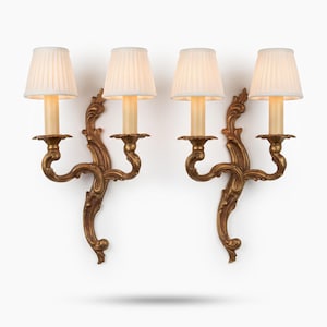 Può includere: Una coppia di applique a due luci con bracci in metallo dorato dalle volute decorate e placche centrali. Ogni lampada è completata da due paralumi in tessuto plissettato color bianco sporco che diffondono una luce calda.