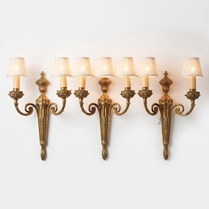 Puede incluir: Tres apliques de pared de metal en tono dorado, cada uno con dos brazos y pantallas de tela plisada blanca. Los apliques tienen una columna central vertical con detalles ornamentados, brazos curvos y acentos florales en la base de cada luz.
