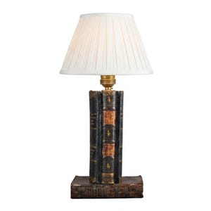 Può includere: Una lampada da tavolo con una base composta da una pila di libri antichi rilegati in pelle con dettagli dorati sul dorso. La lampada include un portalampada color ottone e un paralume in tessuto bianco plissettato di forma conica che emette luce soffusa.