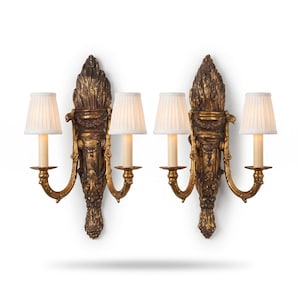 May include: A pair of wall sconces featuring an ornate, dark gold-toned metal backplate with intricate carved detailing and a central face motif. Each sconce has two curved arms supporting white, pleated fabric lampshades and cylindrical bulbs.