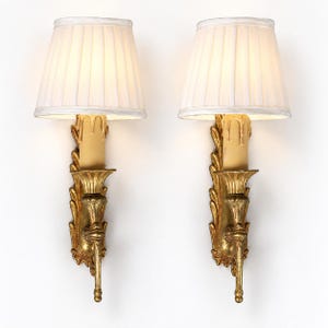 May include: A pair of wall sconces featuring gold-toned metal backplates with ornate leaf detailing and a single candle-style light fixture. Each sconce is topped with a white, pleated, tapered fabric lampshade that diffuses the light.