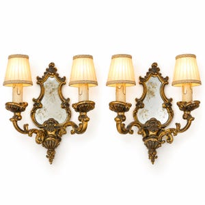 Può includere: Una coppia di applique dorate con cornici in metallo decorate e volute con specchi centrali. Ogni applique presenta due punti luce sormontati da paralumi in tessuto plissettato color crema e copri-candela a goccia.