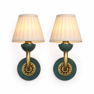 May include: A pair of wall sconces featuring off-white pleated fabric shades. Each lamp has a curved gold-toned arm, a circular green base with a decorative beaded gold ring, and a matching green semi-spherical element supporting the light bulb.