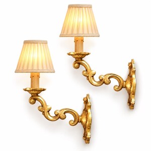 Può includere: Una coppia di applique da parete con bracci in metallo dorato dalle curve ornate e placche decorative. Ogni lampada è sormontata da un paralume in tessuto plissettato di colore chiaro che emette una luce calda e soffusa.