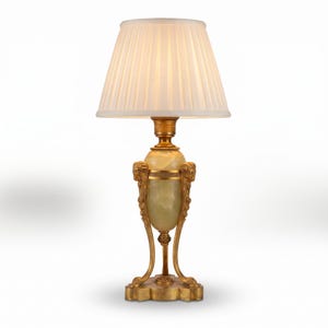 May include: A table lamp featuring a pleated white fabric lampshade atop a central pale green onyx stone body. The base is constructed from gold-toned metal with ornate decorative accents, including sculpted ram heads and curved support legs.