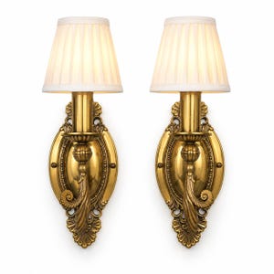 May include: A pair of wall sconces featuring polished brass backplates with ornate scrollwork and beaded detailing. Each fixture includes a single vertical arm supporting a white, pleated, conical fabric lampshade that emits a soft glow.