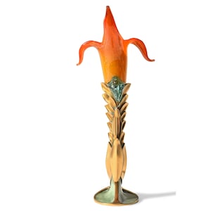 Könnte beinhalten: Eine hohe, orangefarbene Glasvase mit einem goldenen Metallfuß. Die Vase ist wie eine Blume geformt mit einem langen, dünnen Stiel und einer breiten, offenen Öffnung. Der Fuß ist mit einer Reihe überlappender Blätter verziert.
