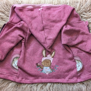 Könnte beinhalten: Eine rosa Kapuzenjacke mit Reißverschluss. Die Jacke hat ein gesticktes Hasenmuster auf dem Rücken mit zwei runden Taschen auf der Vorderseite. Die Jacke hat ein weiches, strukturiertes Aussehen und die Kapuze ist mit einem helleren Rosaton gefüttert.