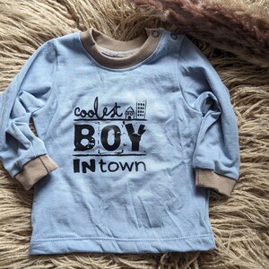 Könnte beinhalten: Ein hellblaues Langarmshirt mit braunem geripptem Kragen und Bündchen. Das Shirt hat einen schwarzen Grafikdruck mit der Aufschrift "coolest BOY in town" mit einem kleinen Haus- und Gebäudedruck.