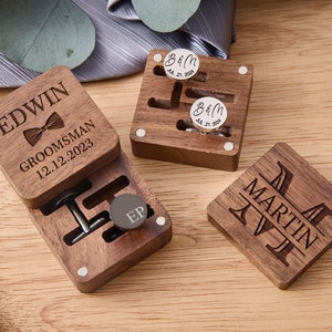 Puede incluir: Tres cajas de madera con mensajes personalizados grabados. Las cajas son cuadradas y tienen un acabado de madera oscura. La caja superior tiene el texto "EDWIN GROOMSMAN 12.12.2023" grabado. La caja del medio tiene un par de gemelos y un broche de corbata en su interior. La caja inferior tiene el texto "MARTIN AVI" grabado.