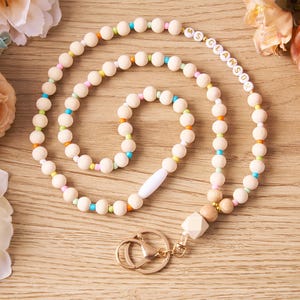 Pode incluir: Um cord&atilde;o de contas de madeira com o nome "MS. CLAWSON" escrito com contas de letras brancas. O cord&atilde;o apresenta uma variedade de contas coloridas e um fecho dourado. O cord&atilde;o &eacute; mostrado em uma superf&iacute;cie de madeira com flores.