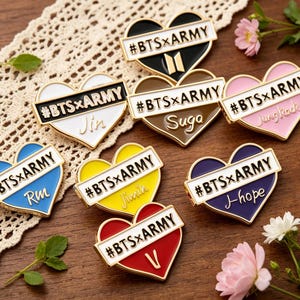 Op de afbeelding: Een verzameling hartvormige emaille pins in verschillende kleuren, waaronder zwart, wit, bruin, roze, blauw, geel en rood. Elke pin heeft de tekst "#BTSxARMY" en een naam of symbool. De pins liggen op een houten oppervlak met kant en bloemen.