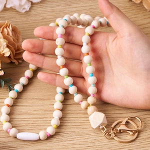 Pode incluir: Um cord&atilde;o de contas de madeira com um pingente geom&eacute;trico branco e ferragens douradas. O cord&atilde;o apresenta contas redondas de madeira de cor clara intercaladas com contas menores e coloridas em tons de rosa, laranja, amarelo, verde e azul.